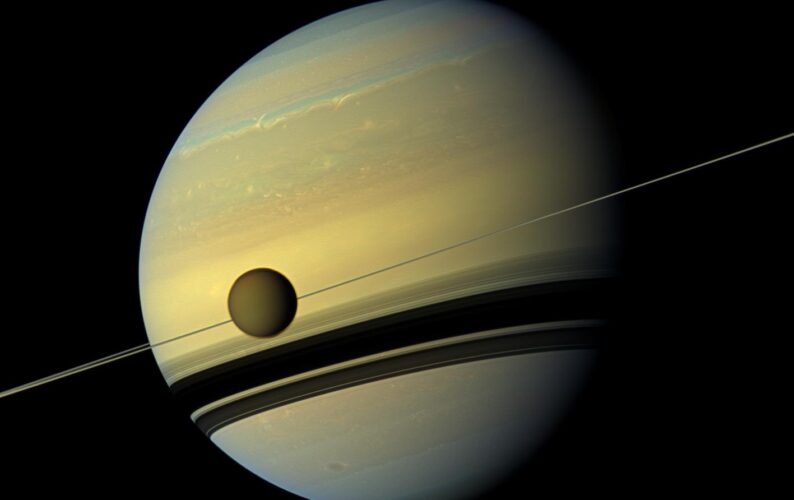 Titan orbits Saturn. Below Titan are the shadows cast by Saturn’s rings.