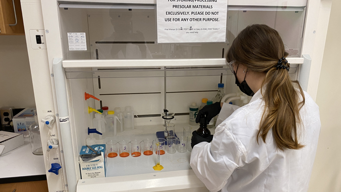 Student wearing lab coat, gloves, goggles, and mask works at a fume hood.