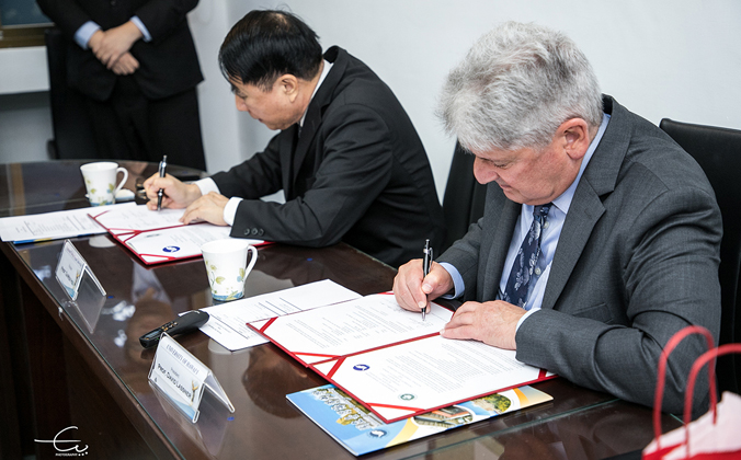 President Xu Taiwen and President David Lassner sign the agreement side by side at a desk. Documents are in red-rimmed folders.