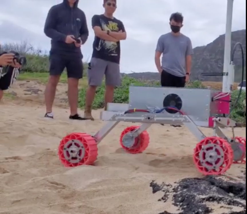Four students test their robotic rover on a beach on O'ahu prior to competition. One students has a camera, another has a remote control device and two others observe.