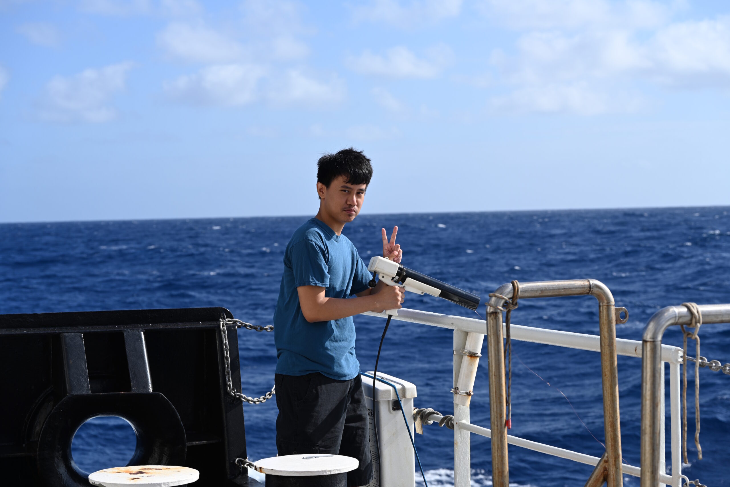 Casey Wandasan aboard the UH research vessel Kilo Moana. He is holding a small instrument with a wire stretching into the ocean and giving a peace sign with the other hand.