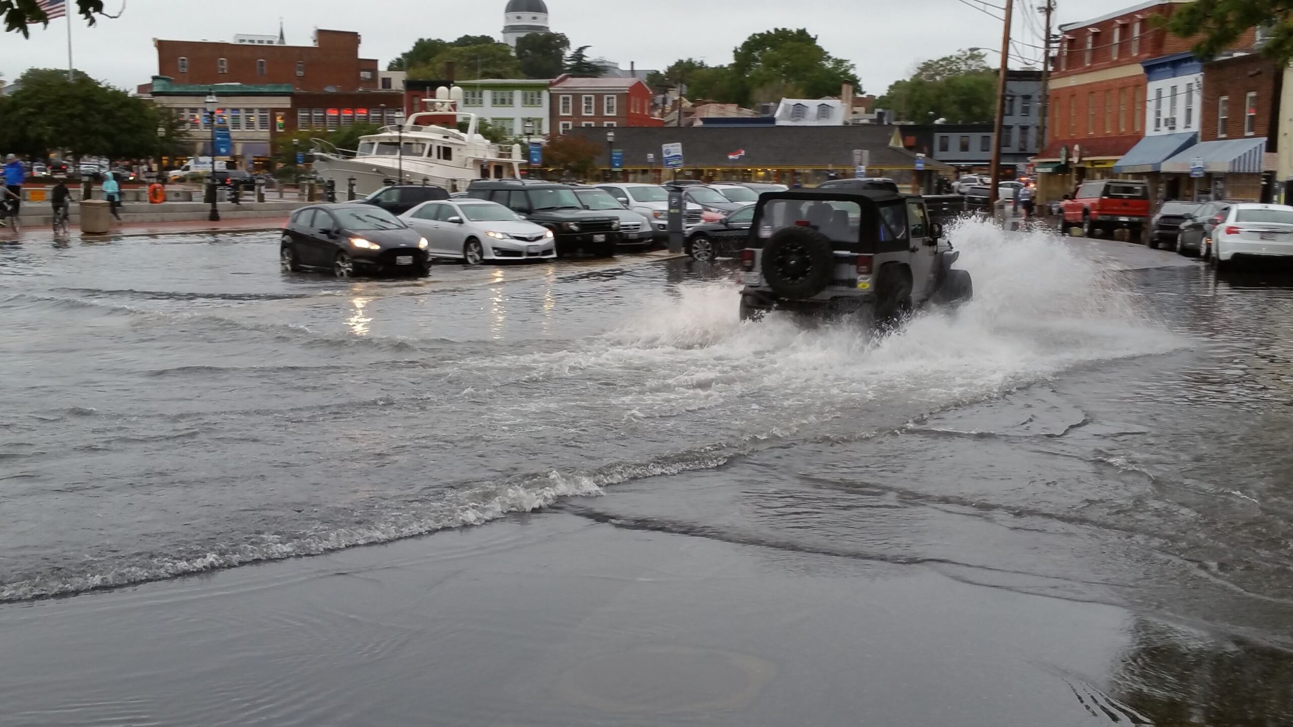 Parked cars line a flooded street as a Jeep drives through, splashing water.