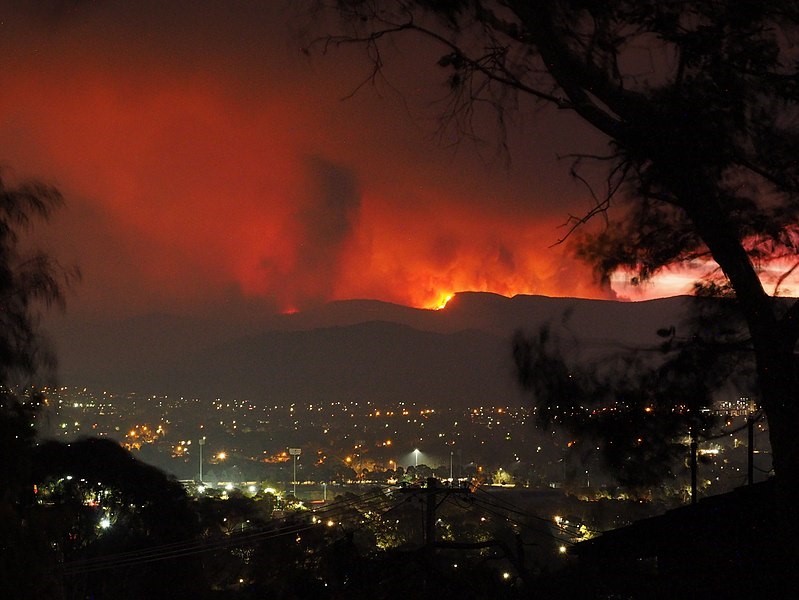 Background: Fire rages behind ridge line; midground: city lights shine in Australia’s capital city, Canberra.