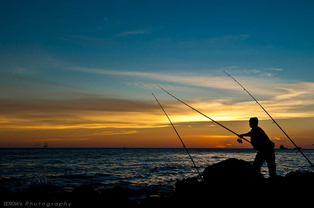 One person is shore fishing with poles while the sun is very low on the horizon.