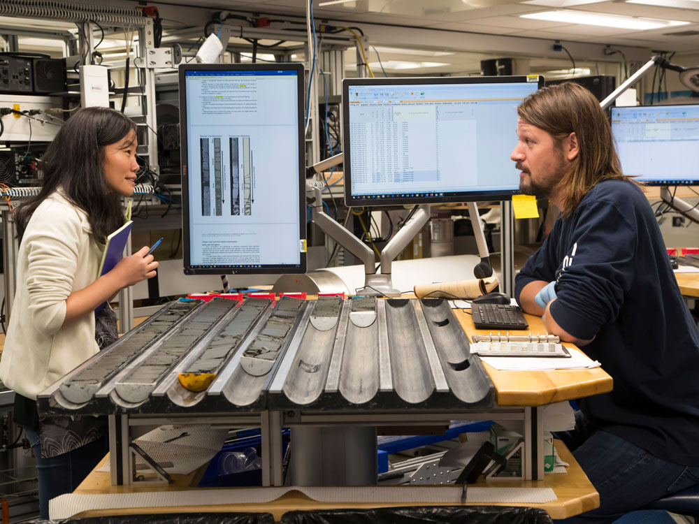 Two researchers stand talking at a table where sediment cores are laid out.
