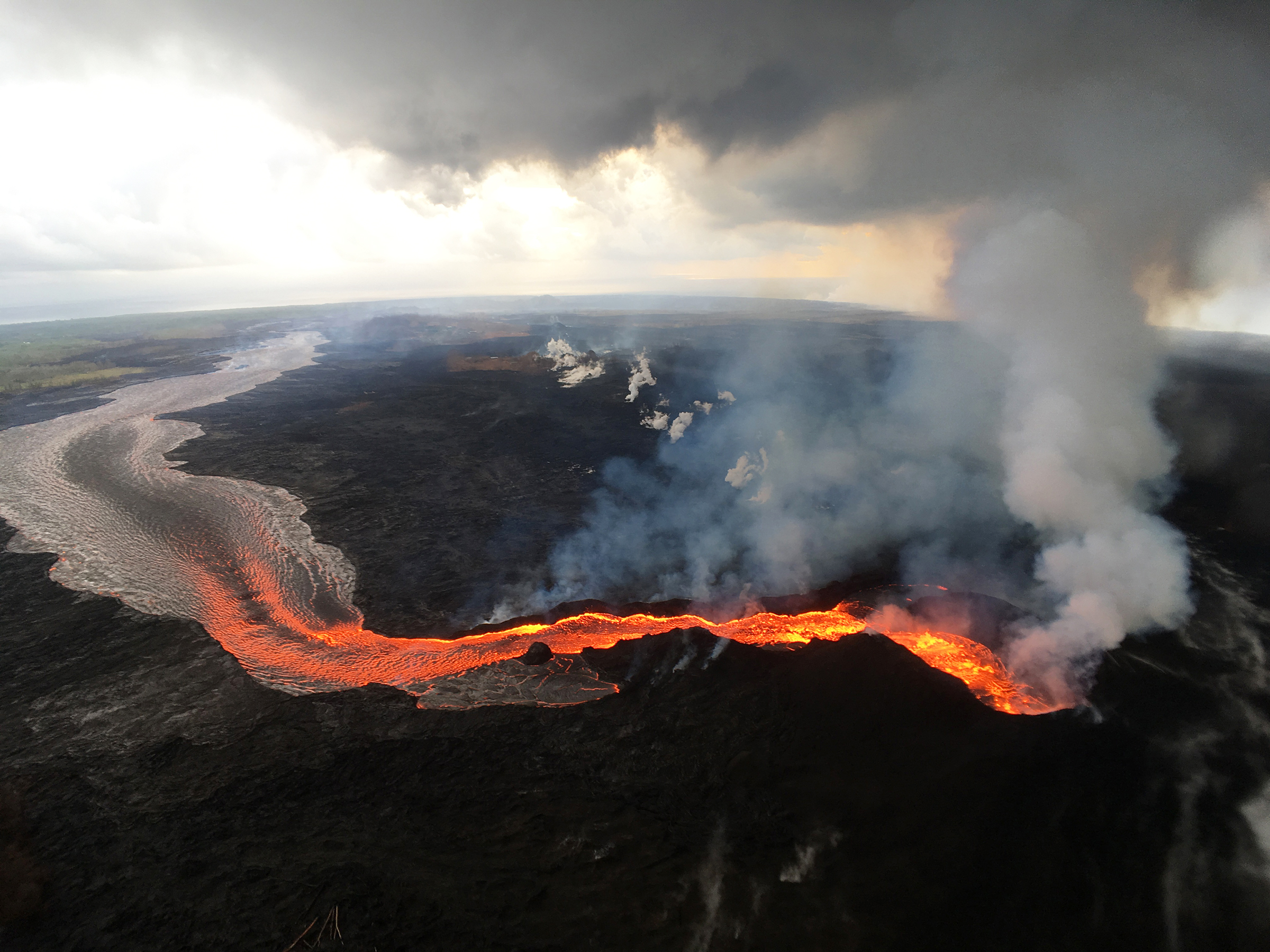 Viewed from above, a river of red bright orange and red lava flows from the site of eruption. White smoke billows high into the sky.