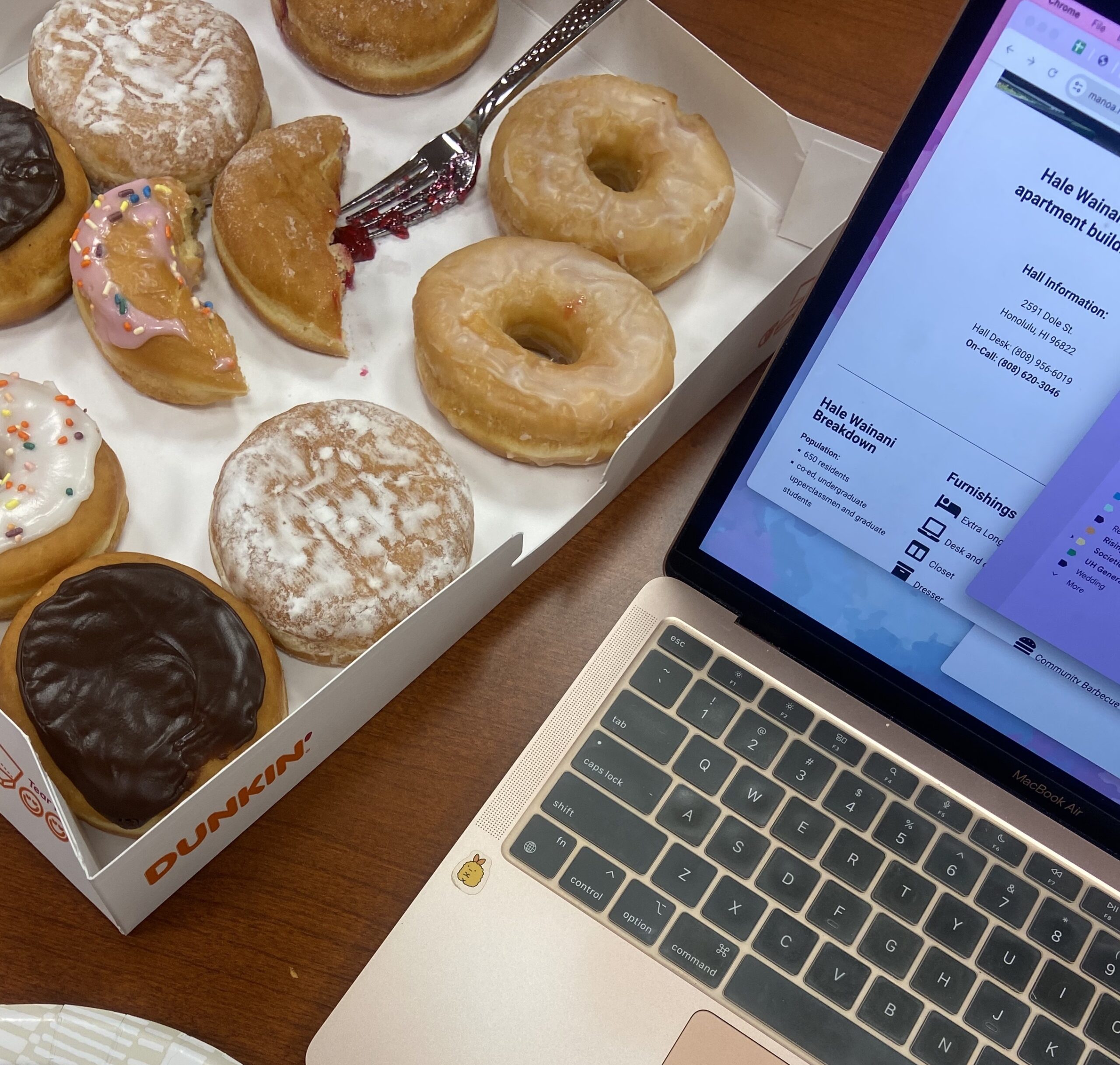 An open box of donuts sits beside an open laptop on a table.