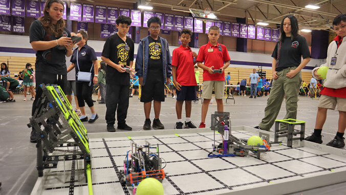 Two student competitors control their robots and four other students and a judge gather around the test platform