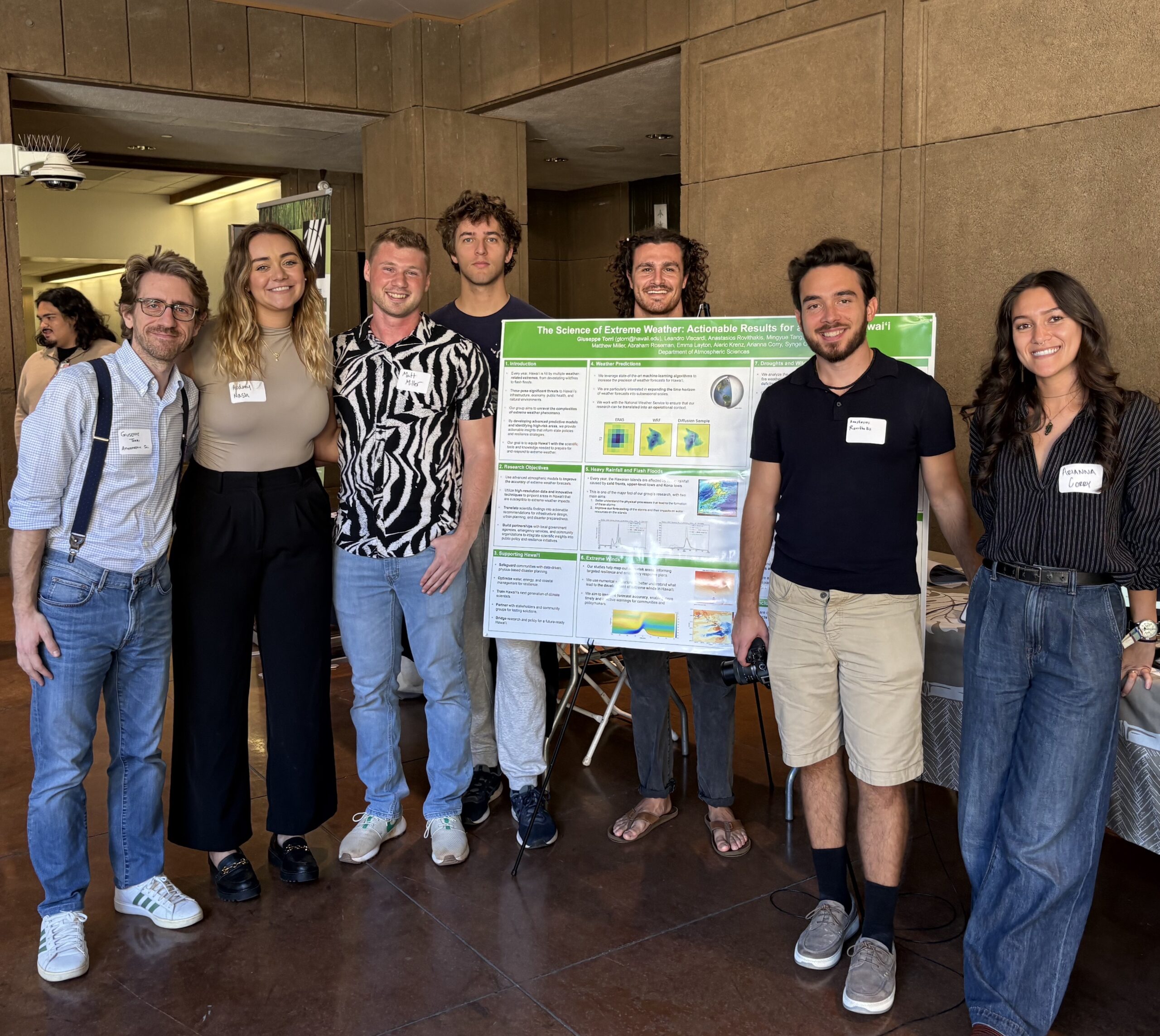 Seven researchers and graduate students gather around a poster and table top display showcasing their atmospheric sciences research