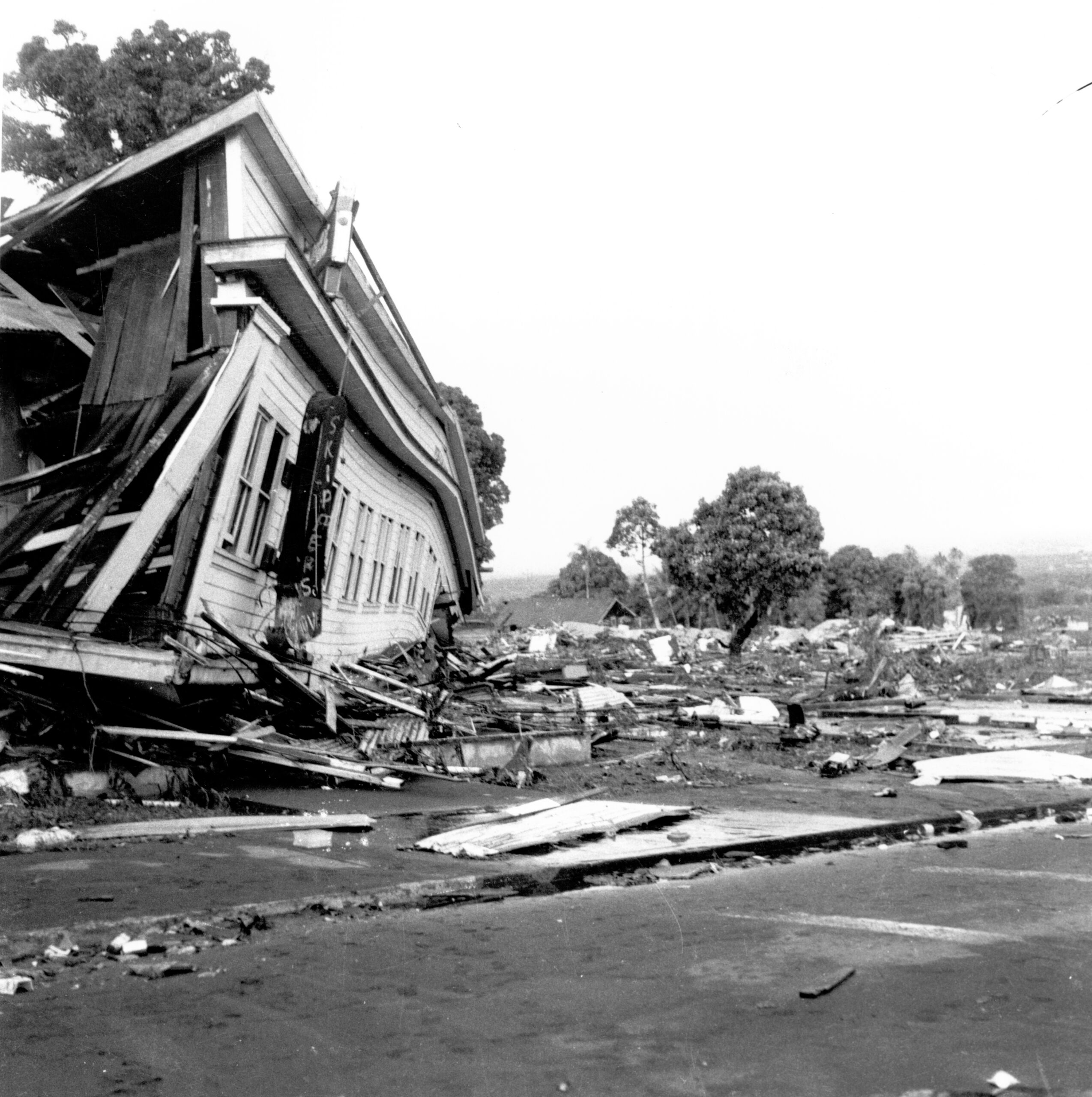 A building is partially collapsed and there is rubble and debris beside the structure. Trees are still standing in the background.