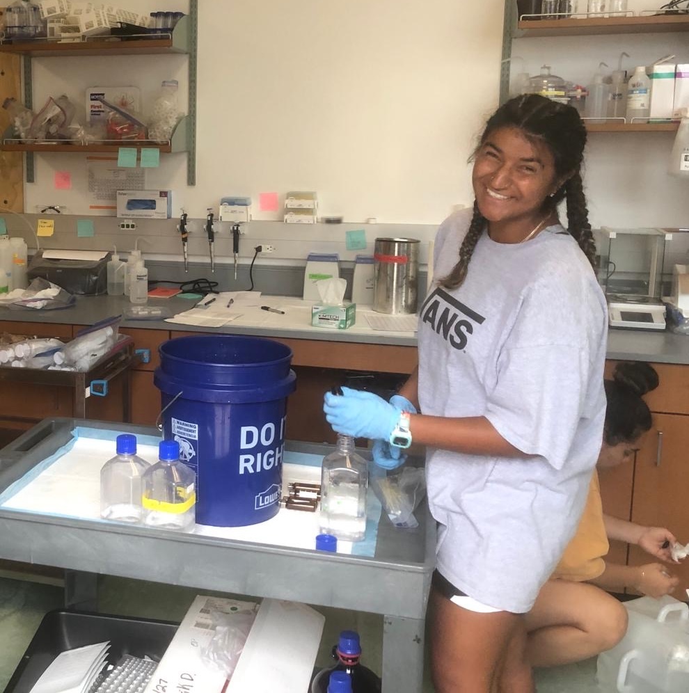 Maya Singh, smiling, works with water samples in clear bottles on a lab cart.